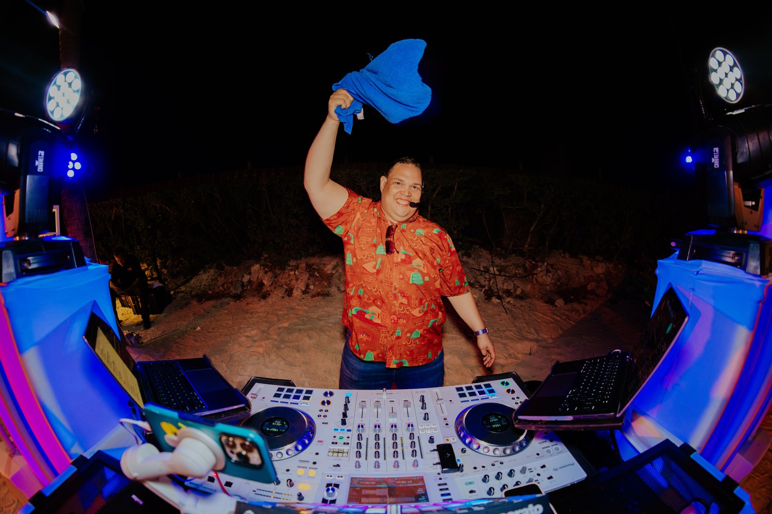 Ruuben smiling and raising a blue towel behind his DJ decks under colorful lights